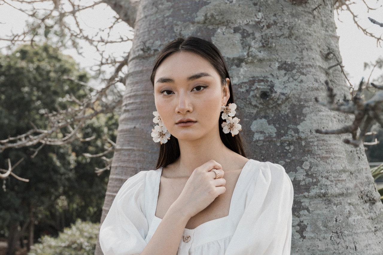 a woman standing in front of a tree in a white dress wearing floral earrings