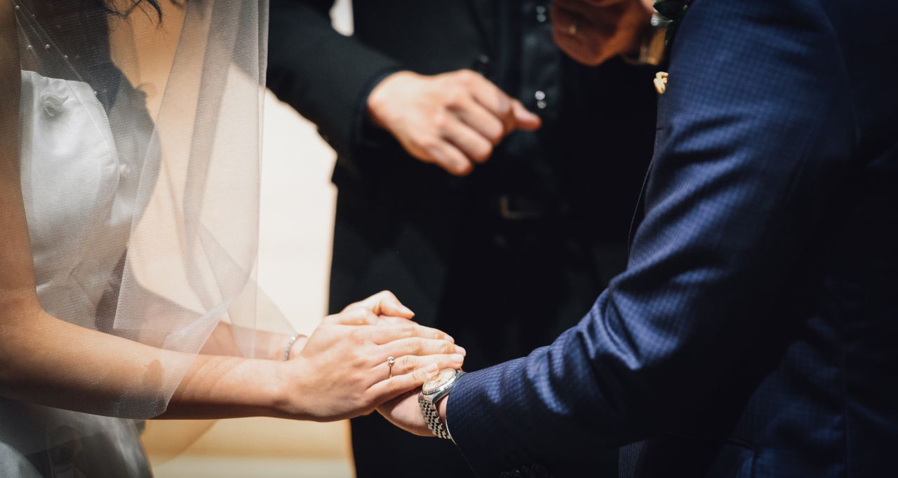 A bride and groom holding hands at the altar, a gold wedding band on the bride’s finger.